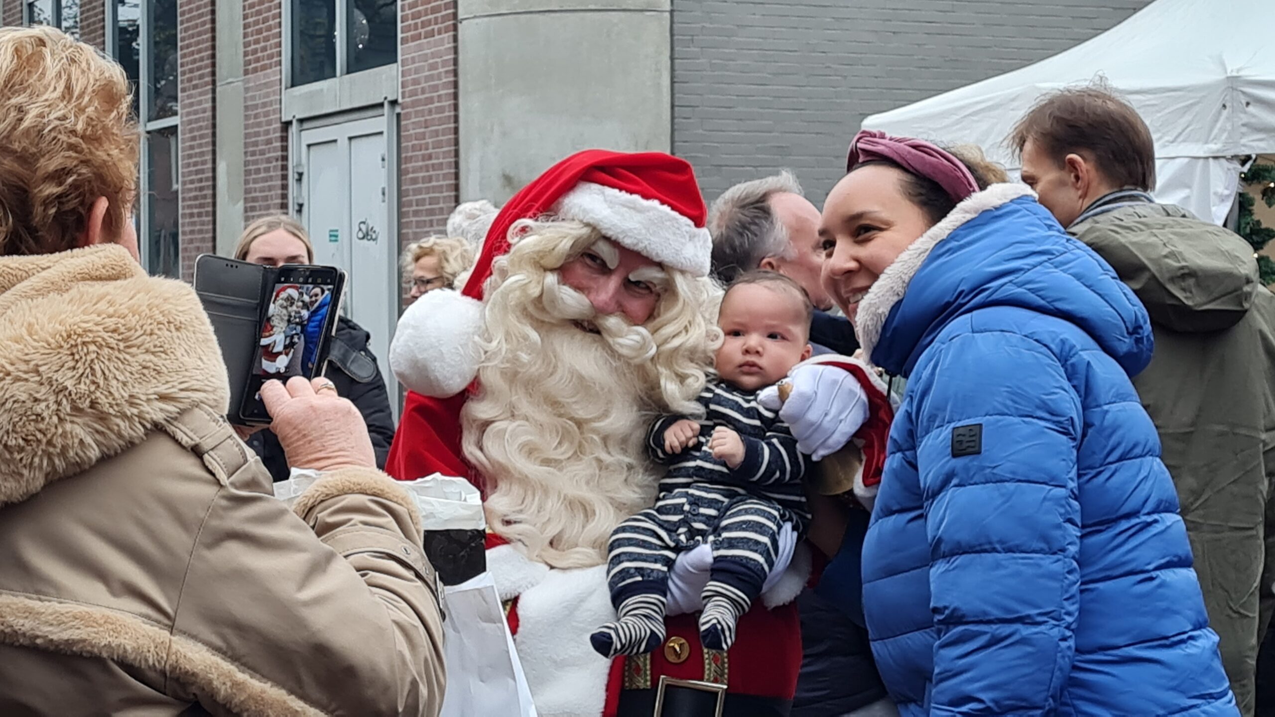 Onze Kerstman kan gitaar spelen en zingen. Dat vinden klanten leuk. Maar ze gaan ook graag met hem op de foto. Hier ze een een moeder met kind en de Kerstman op een drukk winkelstraat.